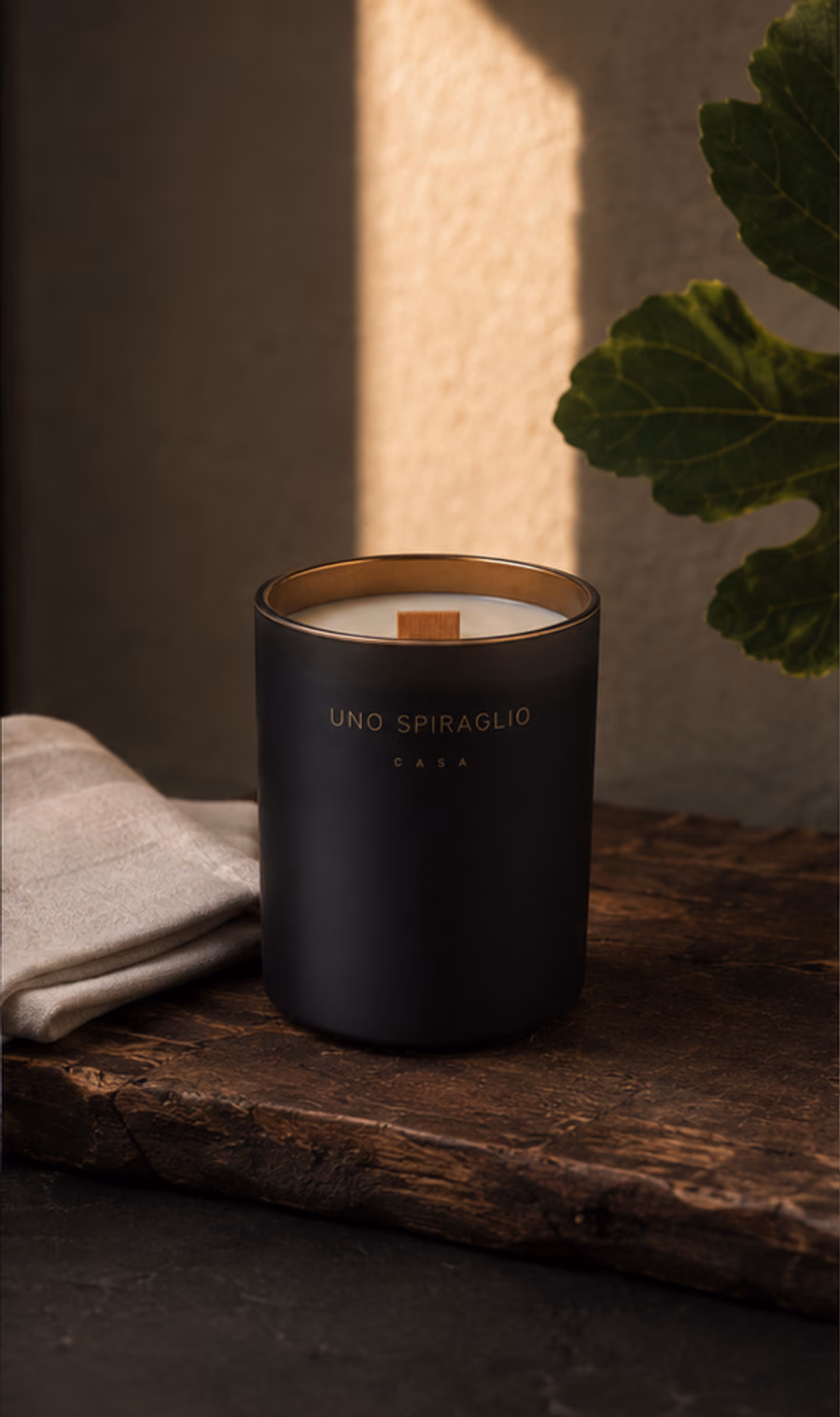La candela Casa su un piano di legno vissuto, con un tovagliolo di lino e una foglia di fico nella luce del pomeriggio.