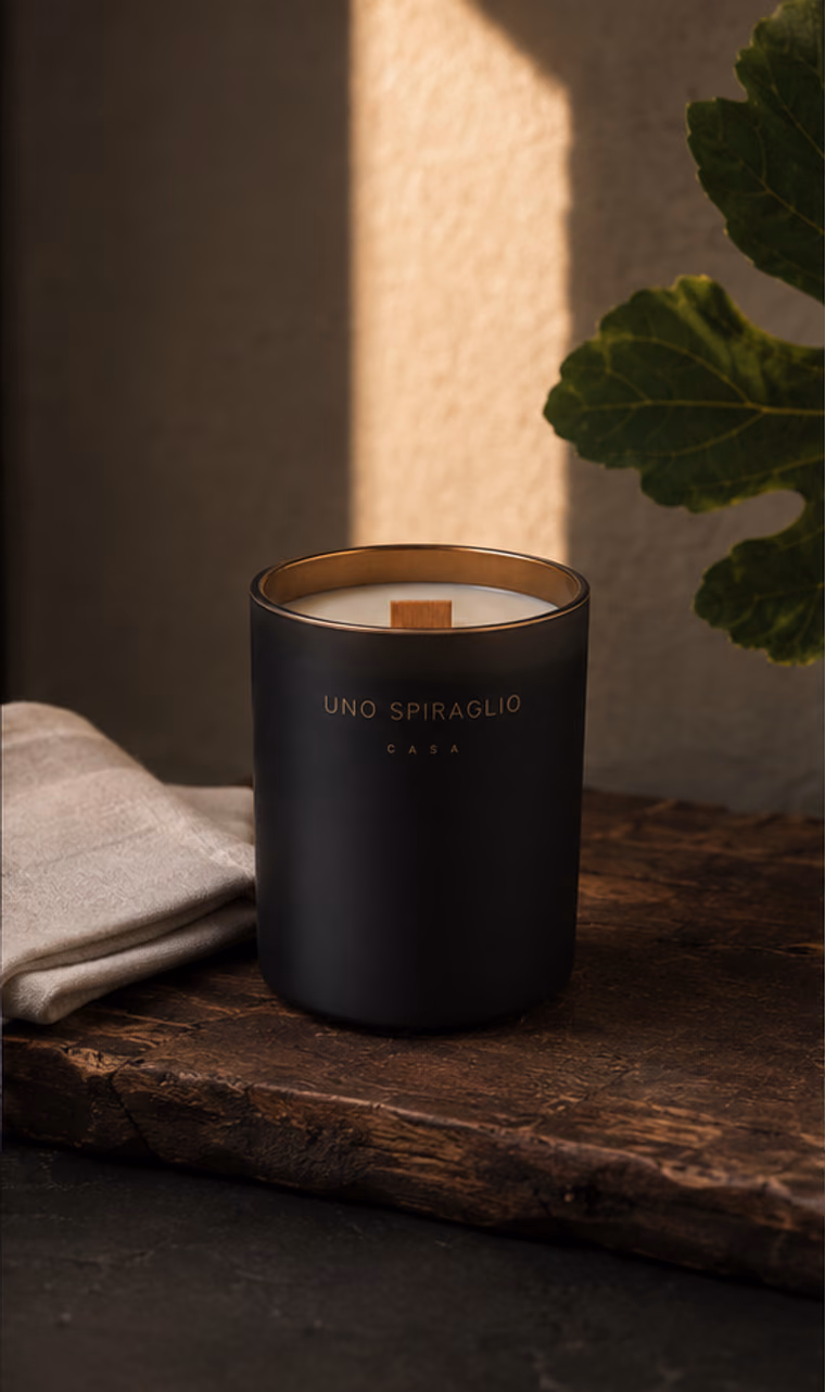 La candela Casa su un piano di legno vissuto, con un tovagliolo di lino e una foglia di fico nella luce del pomeriggio.